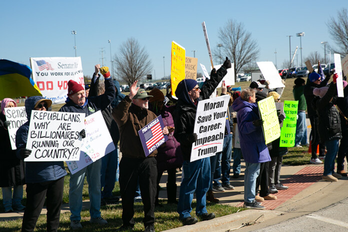 Demonstrators wave signs and hands at drivers who honk in support of their protest against U.S. Sen. Jerry Moran, President Donald Trump and Elon Musk. Photo credit Juliana Garcia.