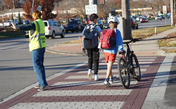 A crossing guard helps two boys, one walking a bike, cross the street.