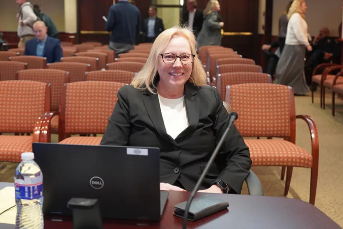 A woman sits behind a desk. Rows of chairs are behind her.