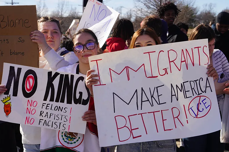 Two people hold up signs that read "NO KINGS" and "IMMIGRANTS MAKE AMERICA BETTER"
