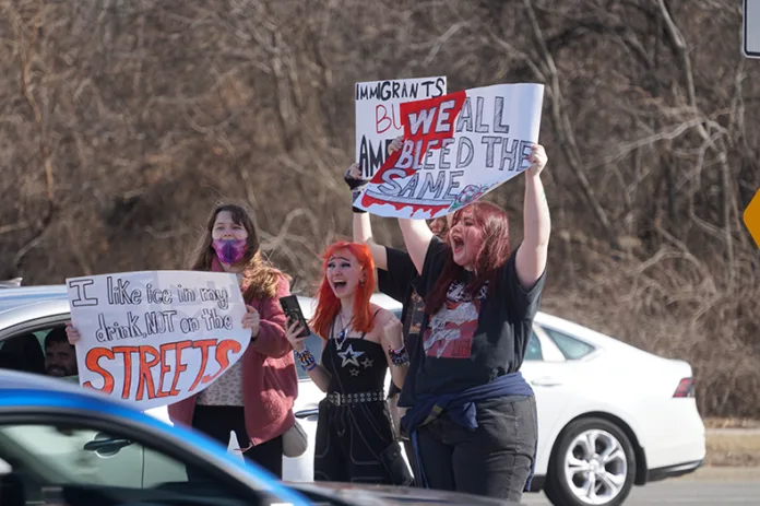 Students hold up signs. Cars pass in the foreground and background.