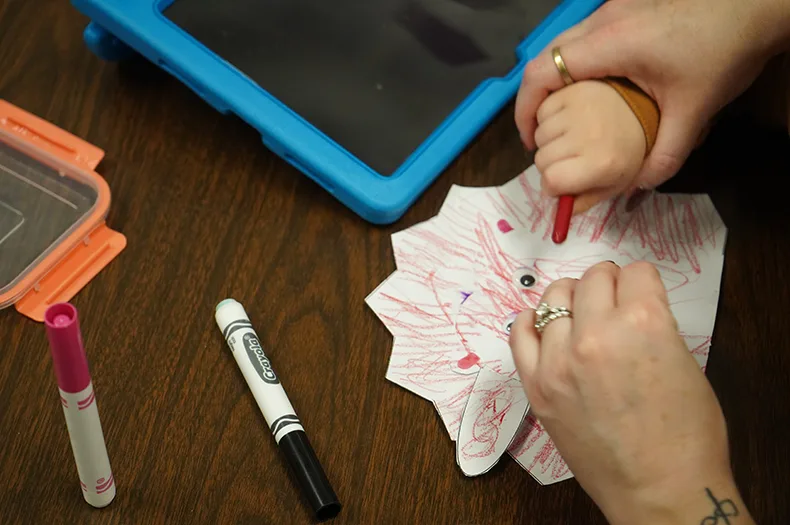 A woman guides a child's hand as he draws.
